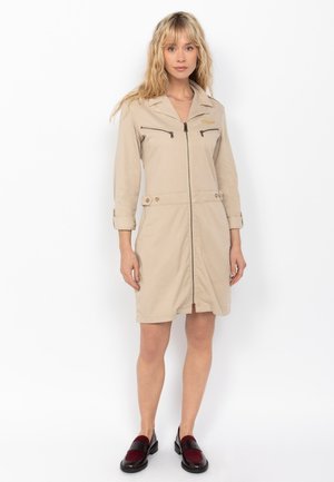 Beige Reißverschlusskleid mit Kragen, Fronttaschen und Knopfdetails. Glattes, leichtes Material. Das Model trägt dunkelrote und schwarze Schuhe.