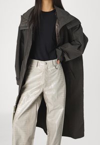 Manteau long vert foncé avec une silhouette structurée, associé à un pantalon gris clair brillant au motif crocodile et une chemise noire en dessous.