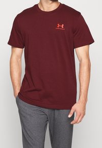 Bordová tričko s krátkým rukávem z bavlny, na hrudi s oranžovým logem Under Armour. Spárováno se šedými kalhotami.