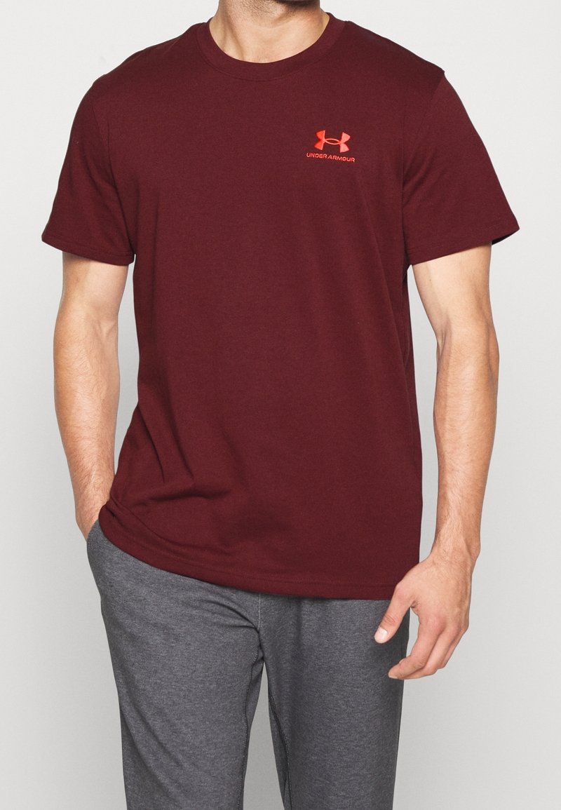 Bordová tričko s krátkým rukávem z bavlny, na hrudi s oranžovým logem Under Armour. Spárováno se šedými kalhotami.