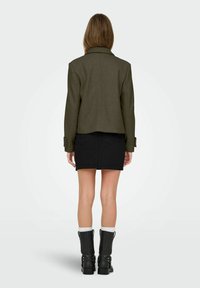 Femme debout de dos portant une veste vert olive, une mini-jupe noire, des chaussettes blanches et des bottes noires mi-mollet sur un fond uni.