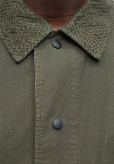 Olívovo zelená bunda s golierom s textúrou rybej kosti, čiernymi gombíkmi a hladkou látkou s viditeľnými švami.