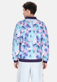  Lichtblauwe bomberjack met een levendig bloemenpatroon van roze en paarse bloemen, groene bladeren, en een donkerpaarse kraag en manchetten.
