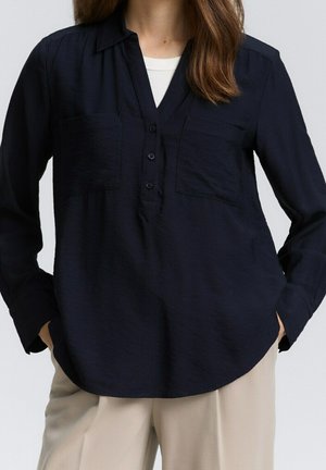 Femme portant une chemise noire à manches longues boutonnée sur un top blanc avec un pantalon beige, mains dans les poches, sur un fond uni.