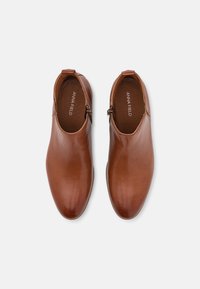 Botas de tobillo de piel marrón con una textura suave, puntera redondeada y cremallera lateral para fácil uso. El interior presenta el nombre de la marca, Anna Field.