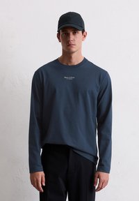 Μακρυμάνικο μπλε πουκάμισο με στρογγυλή λαιμόκοψη, που διαθέτει λευκό σχέδιο λογότυπου στο στήθος; συνδυάζεται με μαύρο παντελόνι και μαύρο καπέλο.