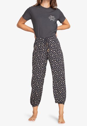 Kobieta w ciemnej koszulce z napisem "Those Summer Days" i luźnych czarnych spodniach z małym beżowym kwiatowym wzorem, stojąca boso z rękami na biodrach.