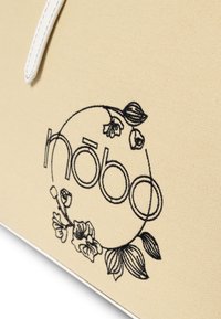 Beige Leinentragetasche mit schwarzem gesticktem Logo und floralen Mustern. Akzente bilden dünne weiße Nähte entlang der Kanten.