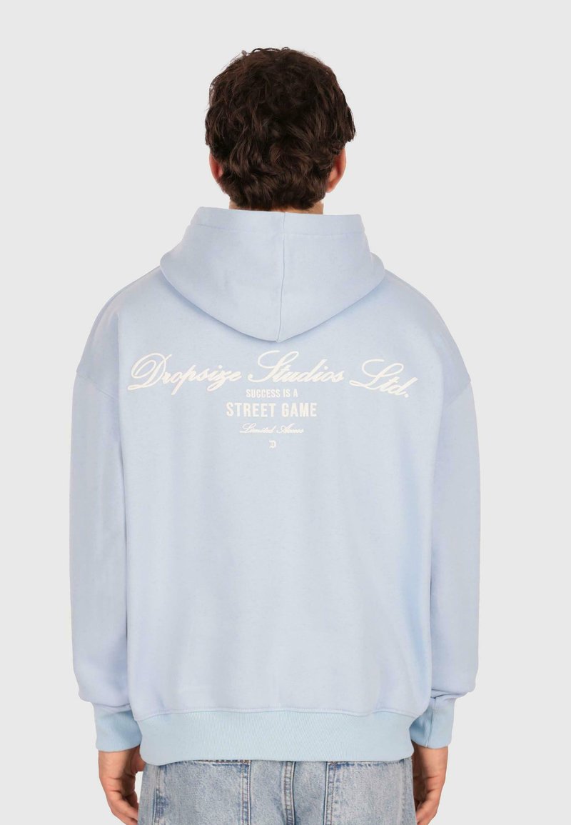 Person, der einen hellblauen Kapuzenpullover mit dem Text "Dropsize Studios Ltd. Erfolg ist ein Straßenspiel" auf dem Rücken trägt, kombiniert mit hellblauen Jeans.