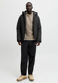 Sort isoleret jakke med hætte, beige strikket sweater, sorte bukser med glans og brune snørebåndsstøvler. Minimalistisk design og teksturer.