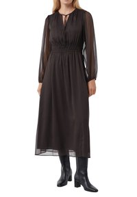 Schwarzes Maxikleid mit langen transparenten Ärmeln, elastischem Taillenbund und Bindeband am Hals. Kombiniert mit knielangen schwarzen Blockabsatzstiefeln.
