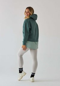 Turkusowa bluza z kapturem, jasna zielona warstwowa bluzka, jasnoszare legginsy oraz kremowe trampki z czarno-białymi paskami na skarpetkach. Gładki materiał i luźny krój.