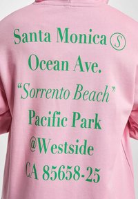 Rosa hettegenser med stor grønn tekst som viser stedsdetaljer: "Santa Monica," "Ocean Ave.," "Sorrento Beach," og en adresse.