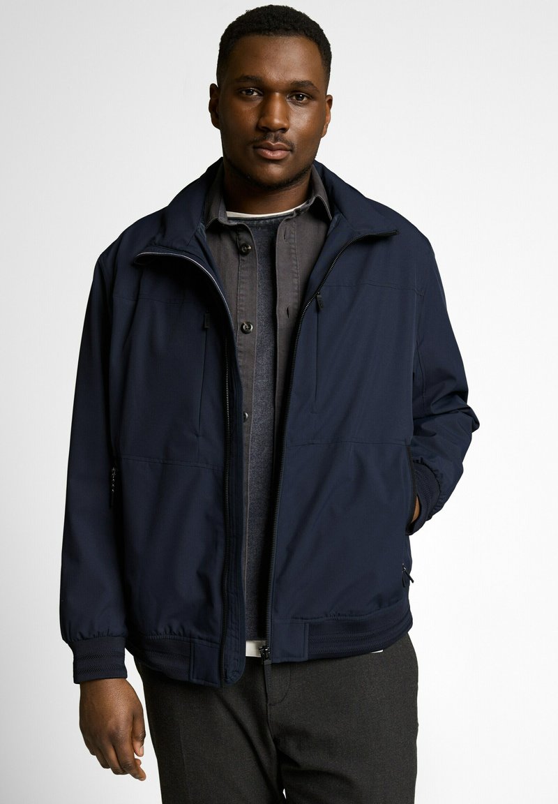 Mladý muž v tmavomodrej bunde na zips cez viacvrstvové tmavosivé tričko a sveter, stojaci na jednoduchom svetlom pozadí.