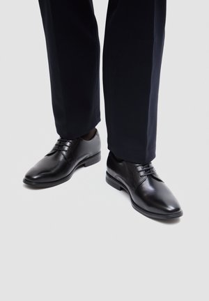 Fekete bőr fűzős elegáns cipők sötét nadrággal, egyszínű világos háttéren.