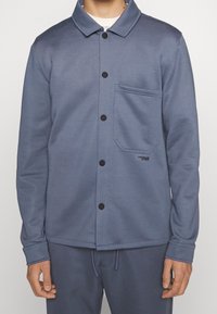 Chaqueta de forro polar azul con cuello camisero, botones negros y un bolsillo en el pecho. Presenta una textura suave y un diseño de ajuste relajado.