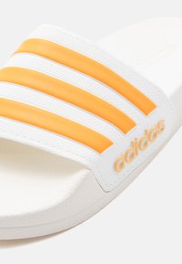 Λευκή σαγιονάρα Adidas με τρεις πορτοκαλί ρίγες στον ιμάντα και το λογότυπο της Adidas στο πλάι.