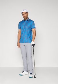 Homme en polo à motifs bleus, pantalon gris clair, chaussures de golf blanches et casquette blanche, tenant un club de golf avec une main gantée, se tenant devant un fond uni.