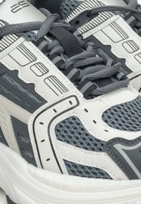 Sportowe sneakersy z teksturowaną białą i szarościową cholewką z akcentami z siateczki, grubą podeszwą i czarnymi detalami w postaci sznurowadeł.
