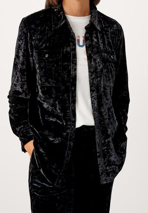 Osoba v černé rozmačkané sametové košili na knoflíky a odpovídajících kalhotách s bílým tričkem s potiskem pod tím. Ruce v kapsách.