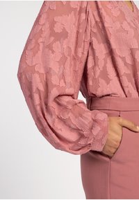 Blouse van doorzichtige roze stof met bloemenborduursel, volumineuze mouwen en een aansluitende tailleband, gecombineerd met een bijpassende roze rok.