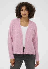 Cardigan rosa chiaro lavorato a maglia con una texture morbida, apertura frontale e orlo a coste. Abbinato a una maglietta bianca e jeans scuri.