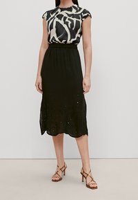 Jupe noire à ourlet festonné avec des détails brodés, associée à un haut à manches courtes avec un motif abstrait noir et beige. Talons marron à brides.