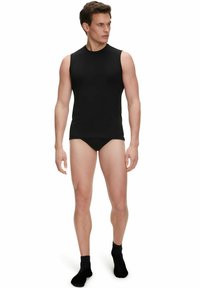 Canottiera nera senza maniche da uomo realizzata in un tessuto morbido, con scollo rotondo e design aderente. Indossata con slip neri e calzini alla caviglia.