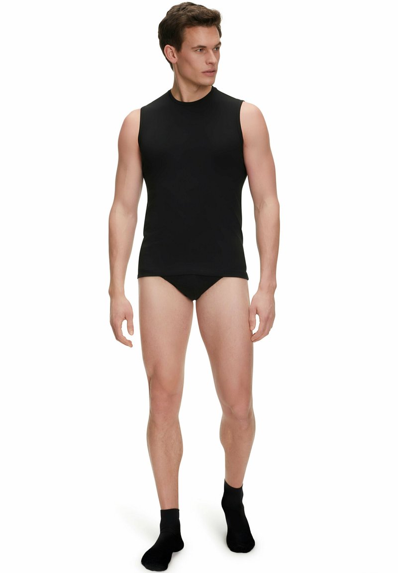 Canottiera nera senza maniche da uomo realizzata in un tessuto morbido, con scollo rotondo e design aderente. Indossata con slip neri e calzini alla caviglia.
