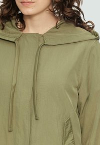 Helle olivgrüne Kapuzenjacke aus einem strukturierten, leichten Stoff. Mit Kordeln am Hals und Seitentaschen. Einfaches Design.