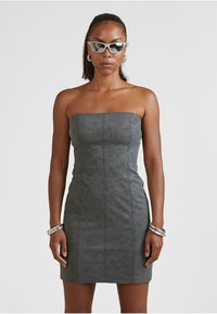 Šedé mini šaty bez ramienok vyrobené z textúrovaného materiálu s vertikálnymi švami, doplnené o angulárne biele slnečné okuliare a strieborné náramky.