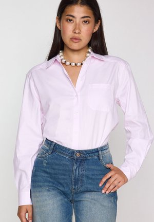 Donna con camicia rosa chiaro abbottonata, jeans di denim e una grande collana di perle, in piedi con una mano sui fianchi.