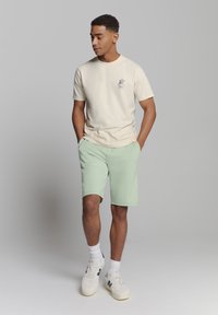 Lichtbeige katoenen t-shirt met een kleine grafische print, gecombineerd met mintgroene shorts. De schoenen zijn witte sneakers met marineblauwe accenten.