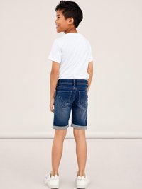 Denim shorts in dark blue with rolled cuffs, paired with a white short-sleeve t-shirt. Model stands with back facing, showcasing the design.