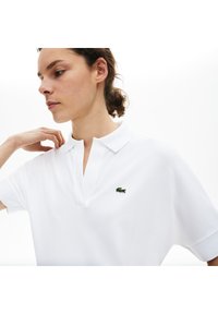Vit kortärmad polo med ribbad krage och en grön krokodil-logo på vänster bröst. Tyget ser slätt och något texturerat ut.