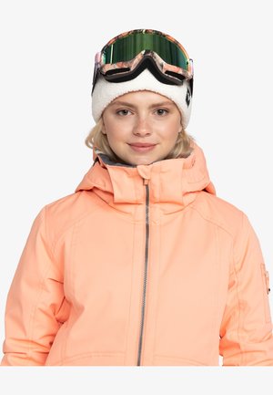 Peach-colored snow jacket with a high collar, zip closure, and side pocket. Model wears a white beanie and green-tinted ski goggles.