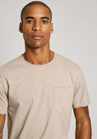 Camiseta de manga corta en color beige claro con cuello redondo y un bolsillo en el pecho izquierdo. La tela tiene un acabado suave y ligeramente texturizado.