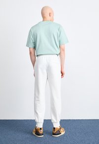 Tricou din bumbac verde deschis, pantaloni cu talie elastică albă, adidași bej și negri, model văzut din spate pe un fundal simplu.