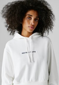 Jonge vrouw met krullend haar, gekleed in een witte hoodie met de tekst "RED BULL RACING" op de voorkant, tegen een effen achtergrond.
