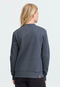 Mørkegrå hettegenser med lange ermer, rund hals, ribbestrikkede mansjetter og nederkant. Har en liten brun lærlogo-patch på nederste venstre side.