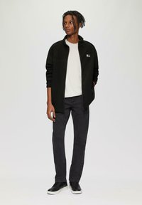 Schwarze Fleece-Zip-Jacke über einem hellgrauen Hemd, kombiniert mit schwarzen Slim-Fit-Jeans und schwarzen Turnschuhen mit weißen Sohlen.
