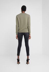 Persona con capelli corti che indossa un felpa verde oliva, jeans skinny neri e scarpe con tacco alte nere, in piedi di fronte a uno sfondo bianco.