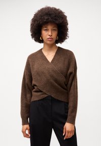 Brązowy sweter zapinany z przodu z długimi rękawami, wykonany z miękkiego, strukturalnego materiału. Wyraźny design z krzyżującym się przodem oraz ściągacze na końcach rękawów. Połączony z czarnymi spodniami.