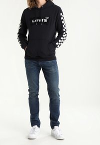 Sweat à capuche noir avec manches à carreaux blancs et logo. Associé à un jean bleu et des baskets blanches, avec une coupe décontractée.