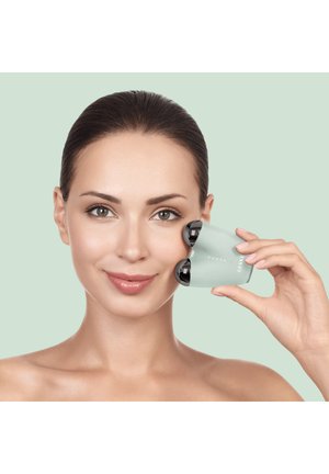 Femme aux cheveux plaqués en arrière utilise un appareil de soin de la peau à double tête sur sa joue droite sur un fond vert clair uni.