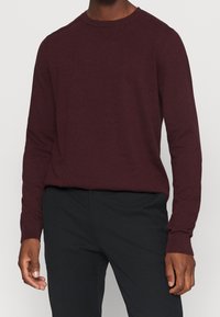 Man i en långärmad, vinröd stickad tröja och svarta byxor, med händerna avslappnat vid sidorna.