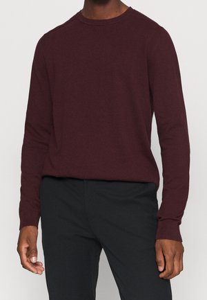 Mann in einem langen, burgunderfarbenen Strickpullover und schwarzen Hosen, mit entspannten Händen an den Seiten.