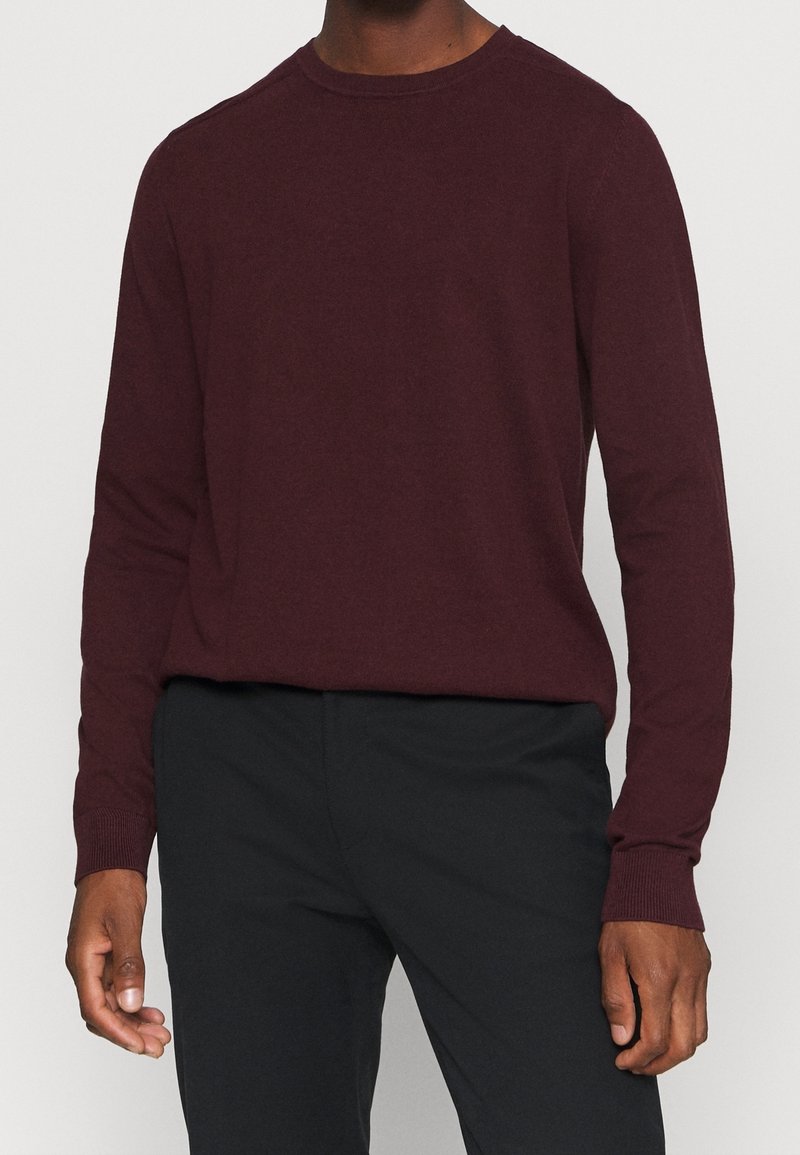 Man i en långärmad, vinröd stickad tröja och svarta byxor, med händerna avslappnat vid sidorna.