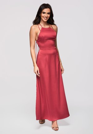 Femme portant une robe de soirée en satin cramoisi sans manches avec des bretelles fines et des côtés ouverts, souriante, debout sur un fond clair uni.