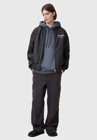 Giacca bomber nera sopra un hoodie grigio, abbinata a pantaloni cargo scuri. La giacca ha una chiusura con zip e un logo sul petto.